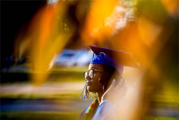   A student with a graduation cap and gown is pictured with orange and yellow autumn tree colors.