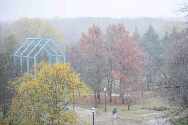  A pedestrian walks through a mix of snowy precipitation on campus filled with autumn tree colors.