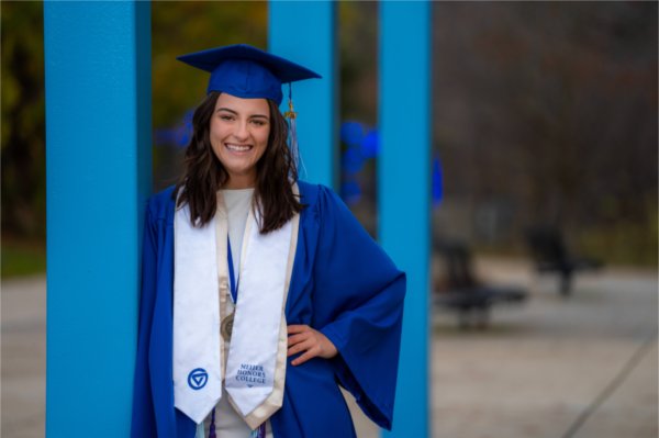 A person wearing graduation garb leans against a blue post.